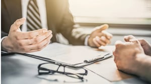 Two individuals in business attire, hands visible as they review documents on a table, with a pair of glasses nearby.